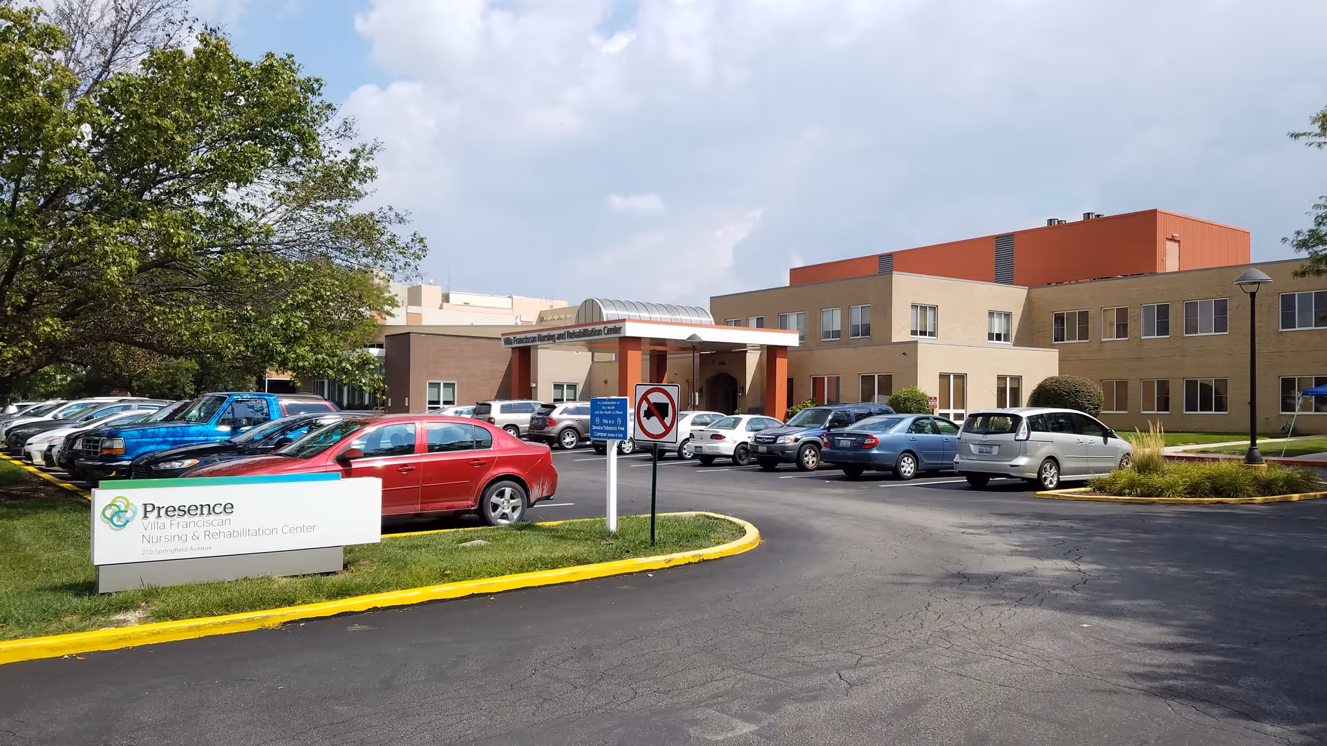 Exterior view of Presence Villa Franciscan Nursing & Rehabilitation Center showing the building, parking lot with several cars, a tree on the left, and a sign with the facility name and address.