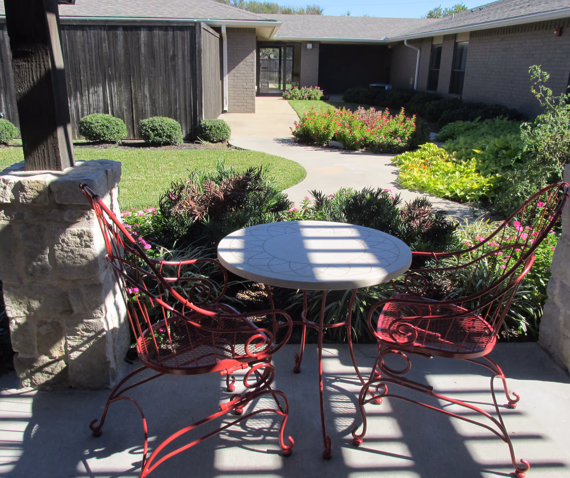 Outdoor patio area with a round table and two red metal chairs under a shaded pergola, surrounded by green bushes and flowering plants, with a walkway and building in the background.