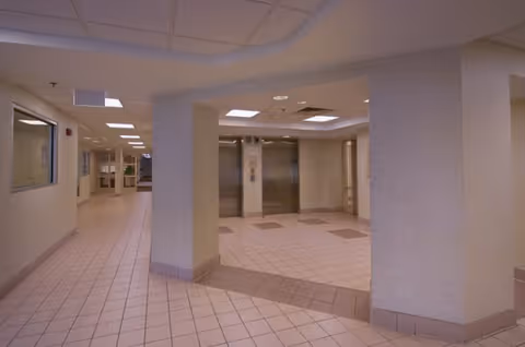 Interior view of a senior living facility hallway with tiled floors and walls. The hallway leads to two elevators with closed doors. The ceiling has recessed lighting and a drop ceiling design. There are windows and doors along the hallway walls.