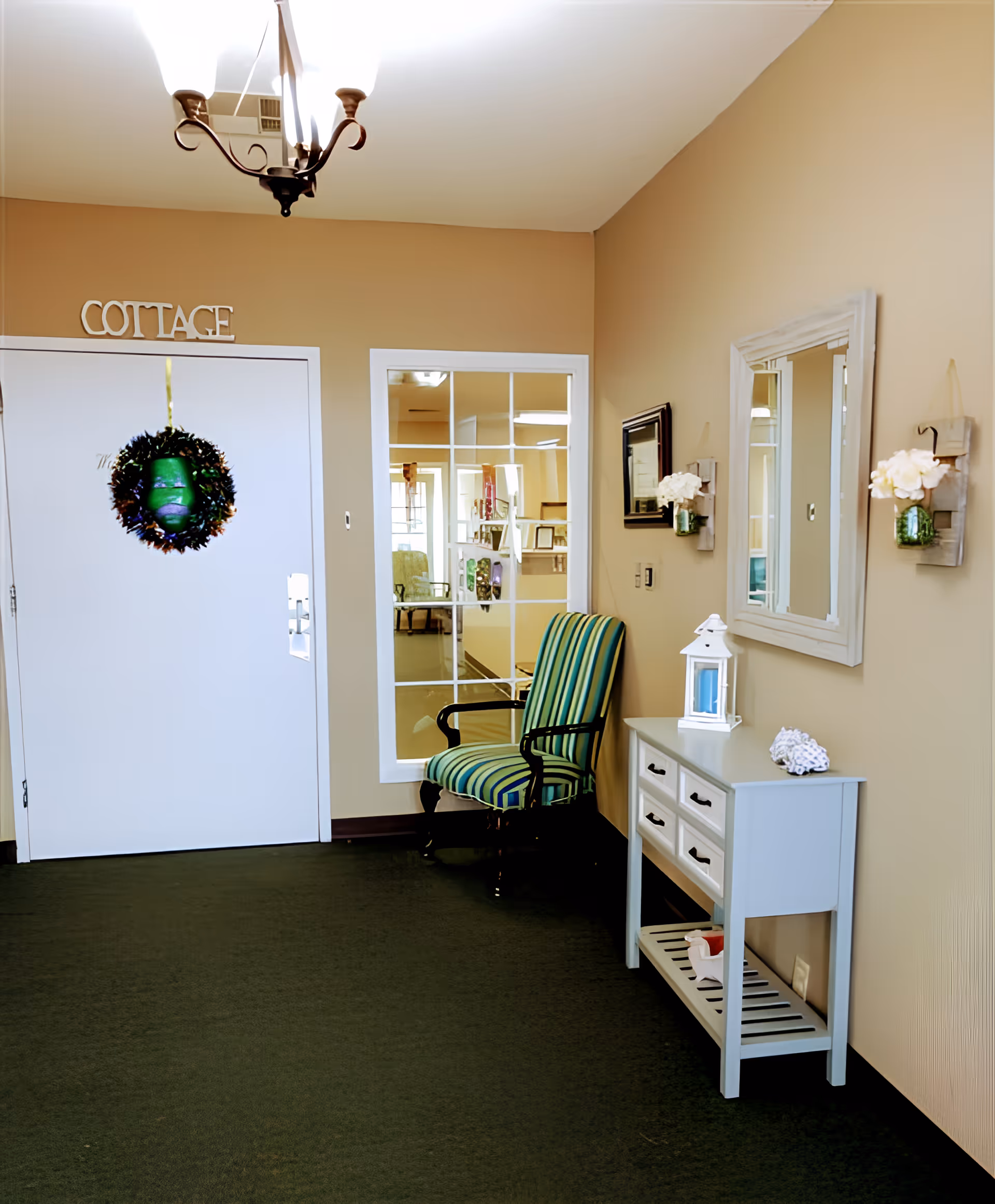 Interior hallway area with beige walls and dark green carpet. A white door labeled 'COTTAGE' has a decorative wreath hanging on it. Next to the door is a large window with white framing. A striped green and blue armchair is placed against the wall. A white console table with drawers and a shelf holds a white lantern and decorative items. Above the table is a white-framed mirror and two small wall-mounted flower holders with white flowers.