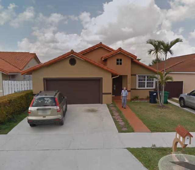 Single-story tan house with red roof and driveway; a man stands near the entrance.