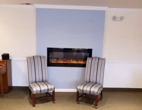 Two striped chairs sit in front of a wall-mounted electric fireplace set in a light blue accent panel in a communal room.