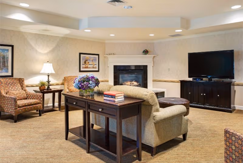 A cozy living room area with a beige sofa, two patterned armchairs, a dark wooden console table with books and a vase of flowers, a fireplace with a white mantel, and a flat-screen TV on a dark wooden cabinet. The walls have light-colored wallpaper with framed artwork, and the floor is carpeted.