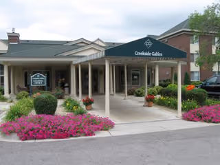 Entrance to Creekside Gables facility with a covered walkway supported by columns, surrounded by well-maintained landscaping including bushes and pink flowers. A sign near the entrance reads 'Creekside Gables Learning Office'.
