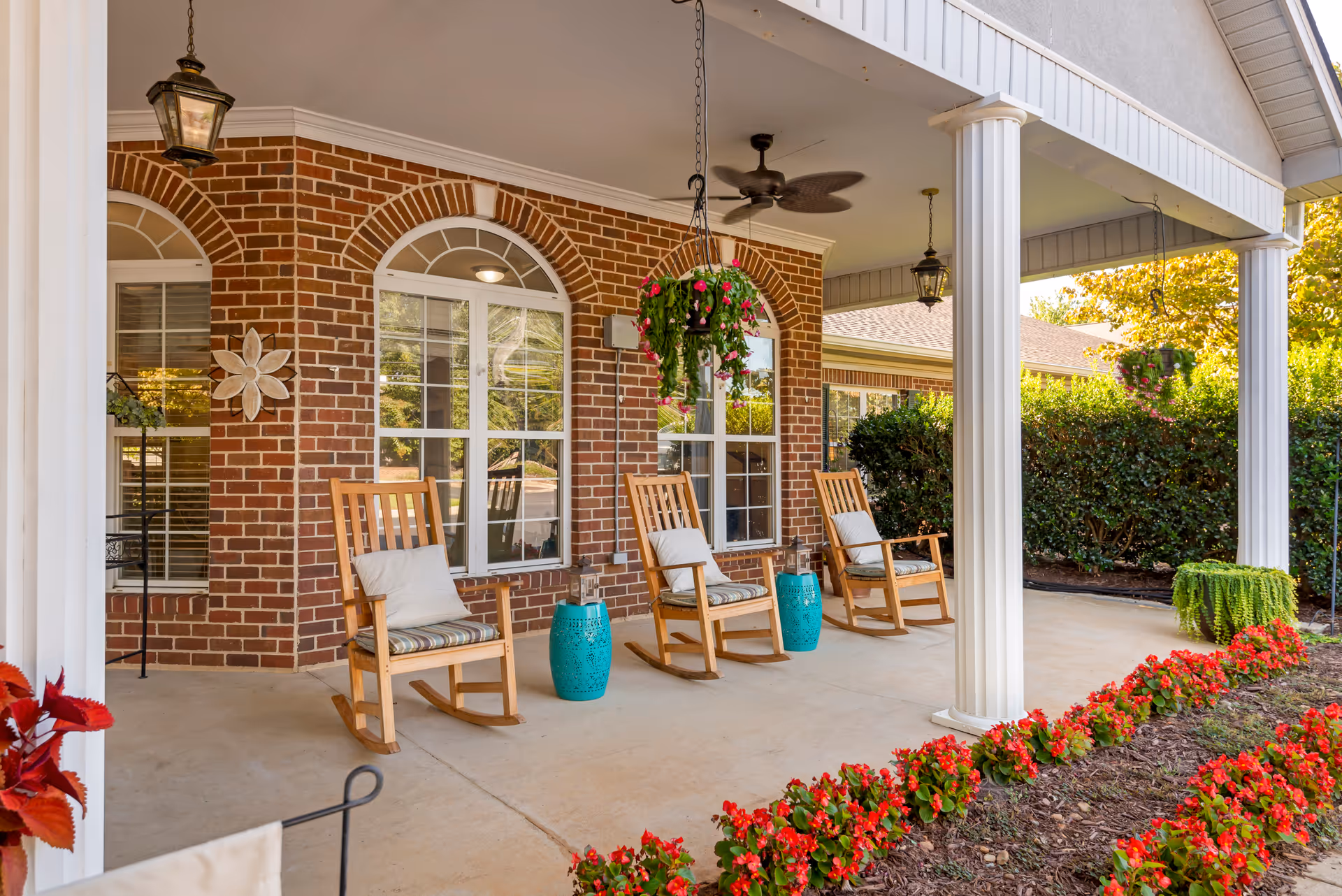 Covered porch area with three wooden rocking chairs each with cushions, two turquoise side tables, hanging flower baskets, ceiling fan, and brick wall with arched windows. Flower beds with red flowers line the edge of the porch.