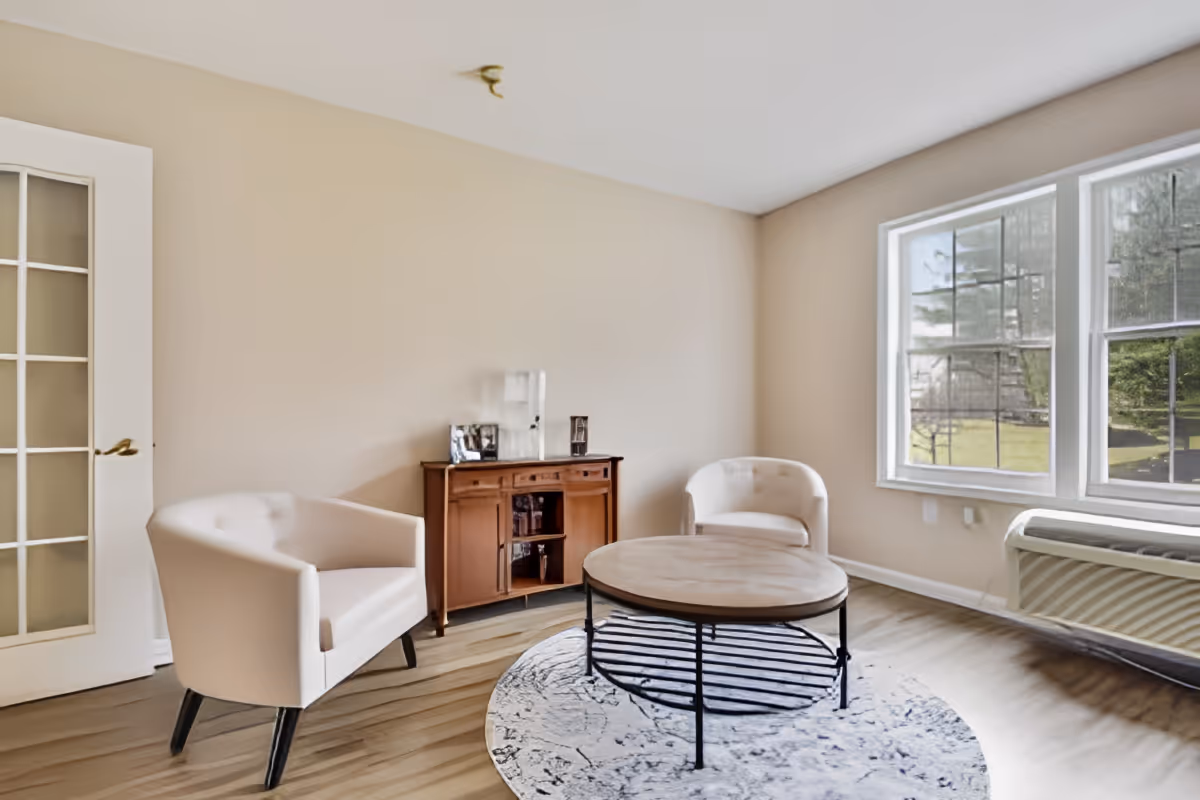 A bright living room with two white armchairs, a round wooden coffee table on a patterned rug, a wooden sideboard with decorative items, large windows letting in natural light, and a glass-paneled door.