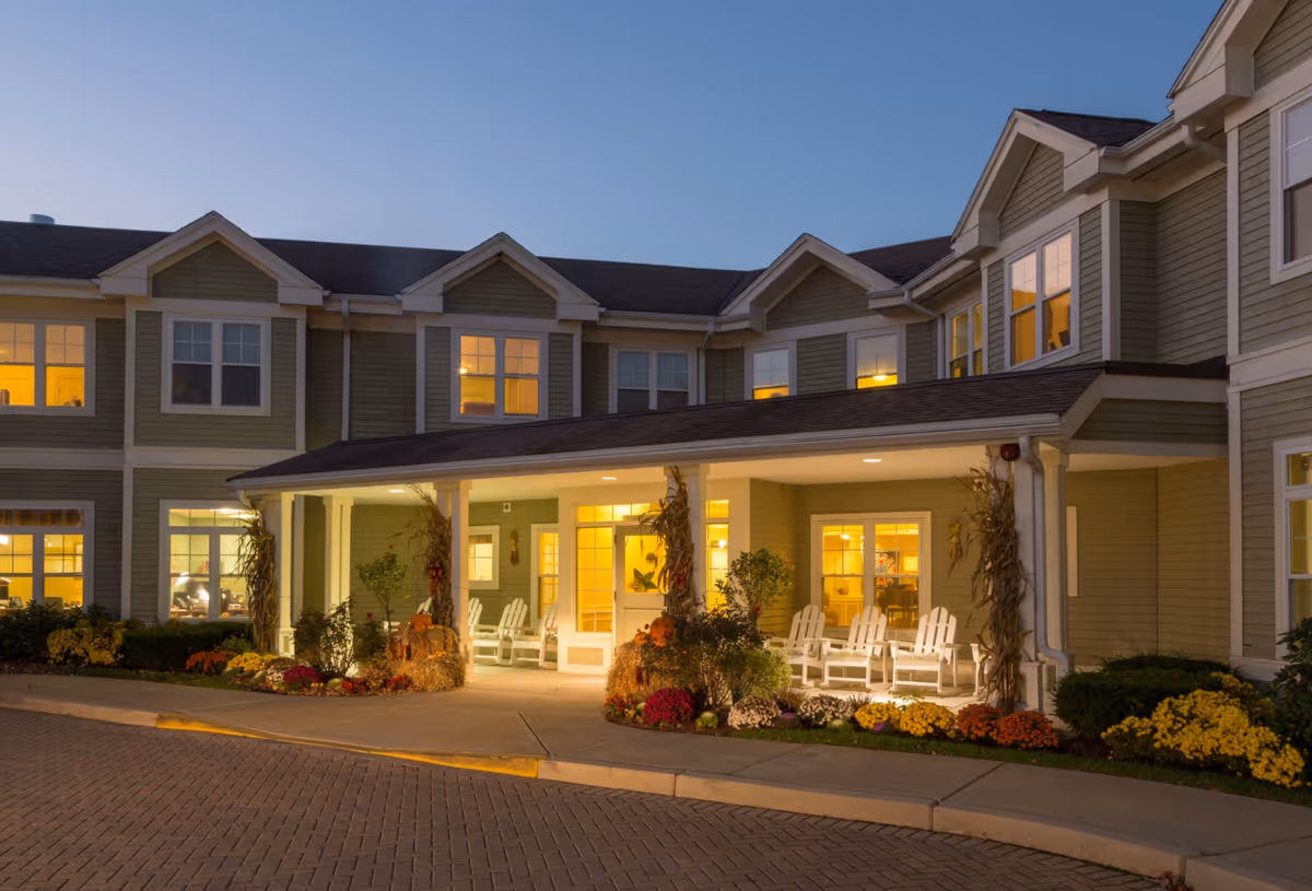 Front entrance of a two-story senior living residence at dusk with a covered porch, white chairs, warm interior lights, and landscaped flower beds.