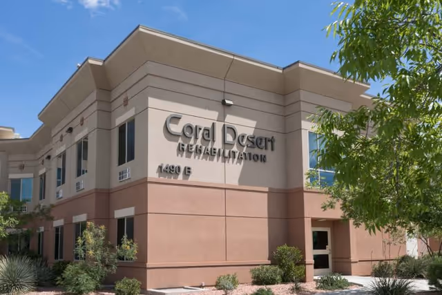 Exterior view of Coral Desert Rehabilitation building with beige and brown walls, several windows, and surrounding desert landscaping including bushes and small trees under a clear blue sky.