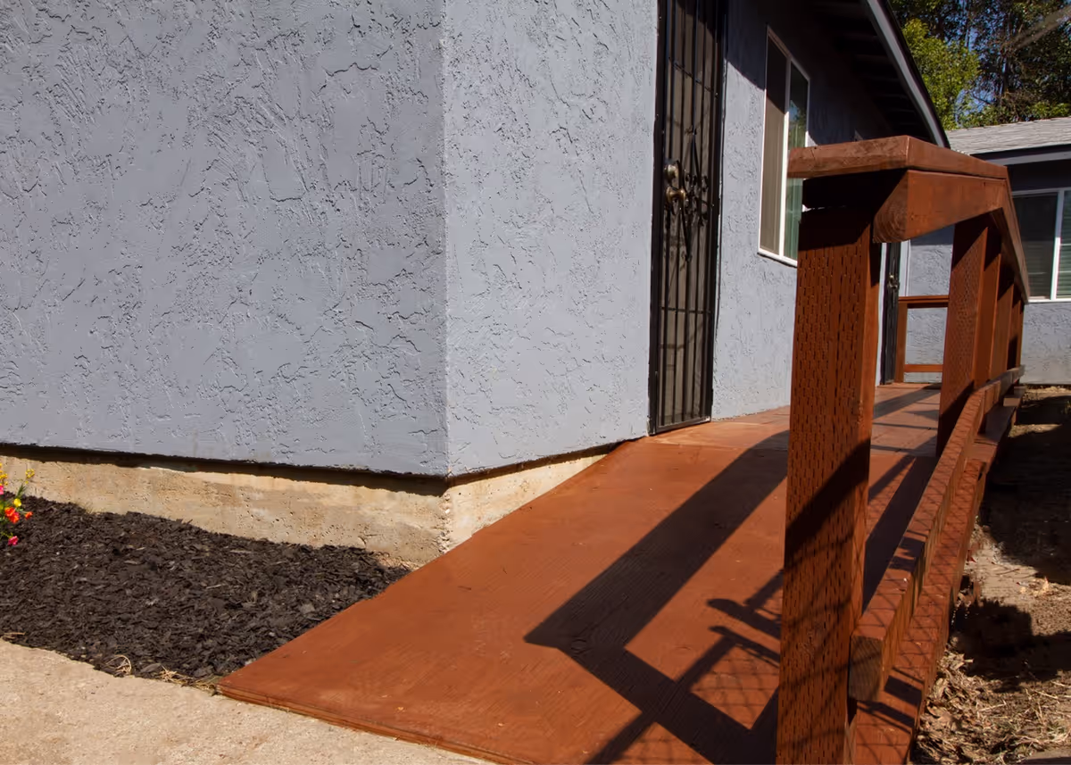 Exterior view of a building corner with a textured gray wall and a brown wooden ramp leading to a black metal door with a window beside it. The ramp has wooden railings on the right side and there is mulch and some small plants on the left side near the building.