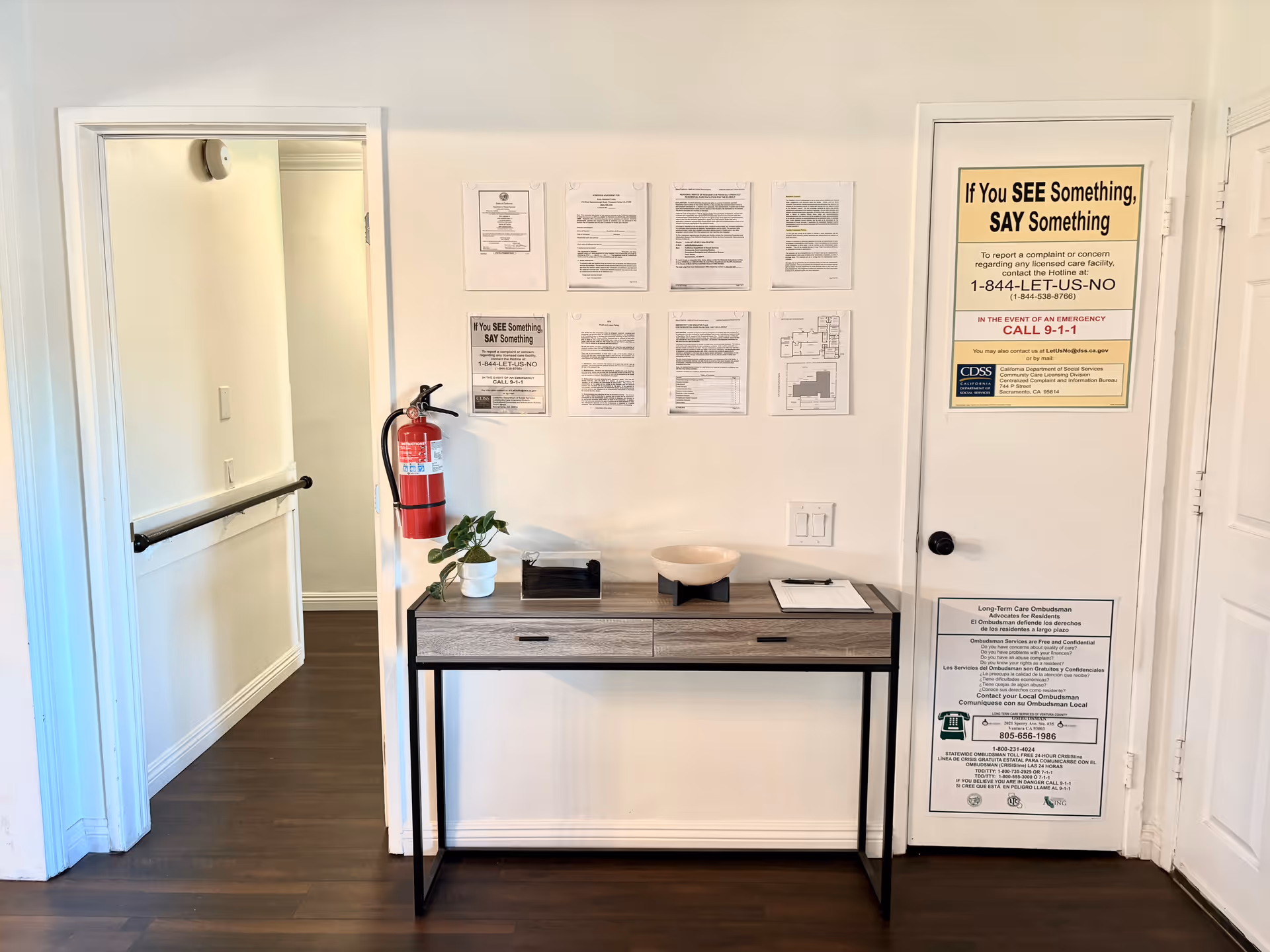 A hallway corner in a senior living facility with a small wooden table against a white wall. On the wall above the table are several notices and informational posters, including a large yellow and black sign that reads 'If You SEE Something, SAY Something.' A fire extinguisher is mounted on the wall to the left of the table. The floor is dark wood, and there is a doorway to the left leading to another hallway with a handrail.