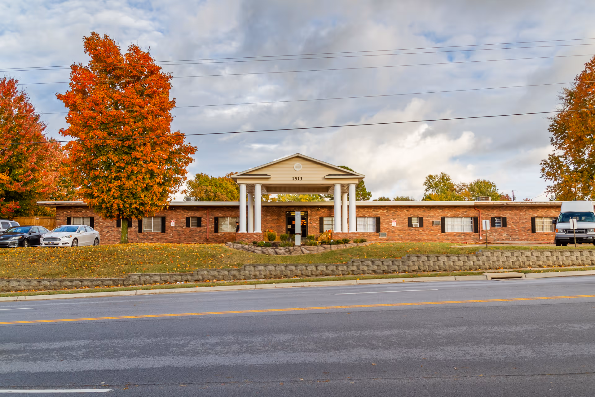 Front exterior view of The Blossoms at Rogers Rehab & Nursing Center, a single-story brick building with a covered entrance supported by white columns. There are autumn-colored trees on either side of the building and several parked cars in front. The sky is partly cloudy.