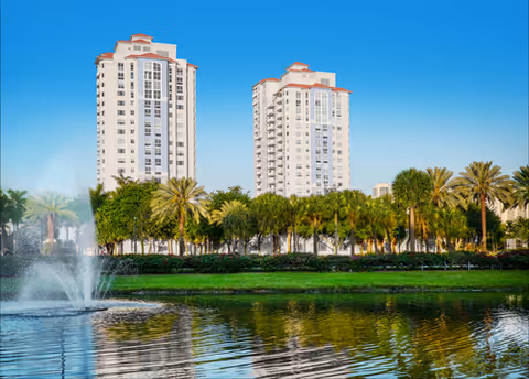 Two white high-rise residential towers behind palm trees with a fountain and reflecting pond in front under a clear blue sky.