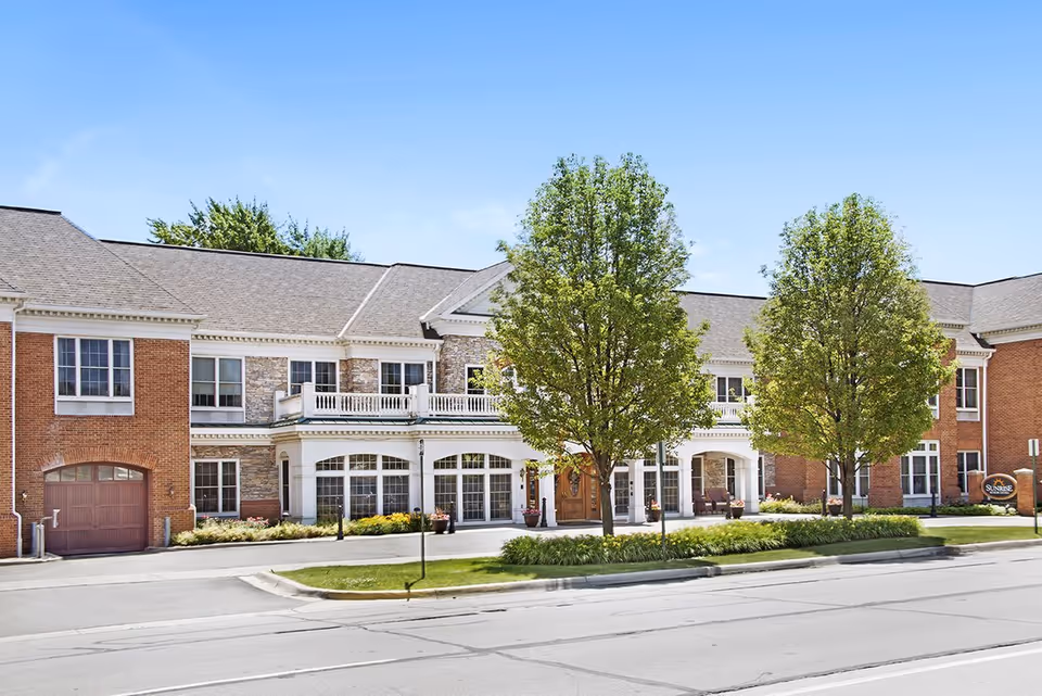 Two-story brick and stone senior living building front with a covered entrance, balcony, landscaped beds and trees.