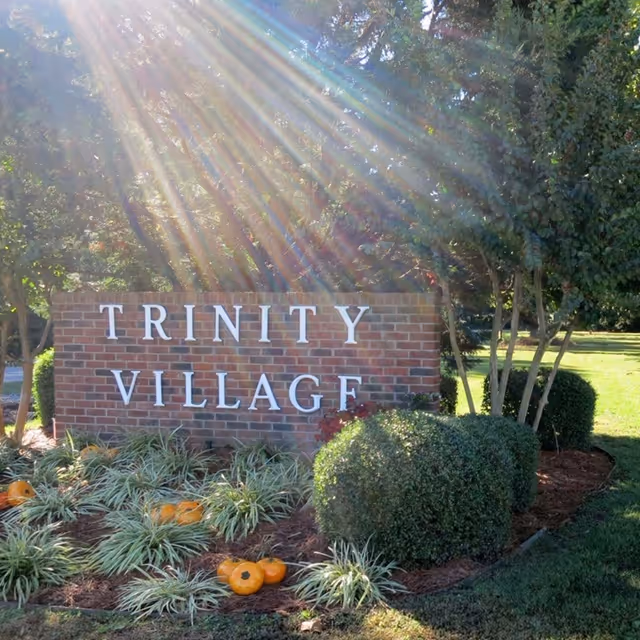 A brick sign with white letters reading 'TRINITY VILLAGE' surrounded by landscaping including bushes, grass, and small pumpkins, with sunlight streaming through trees in the background.