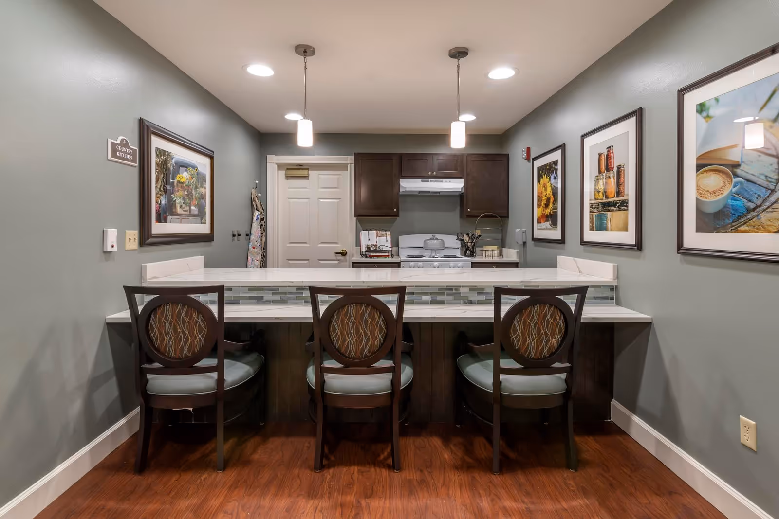 Interior view of a small communal kitchen with a countertop and three chairs facing a stove and cabinets, pendant lights, and framed artwork on the walls.