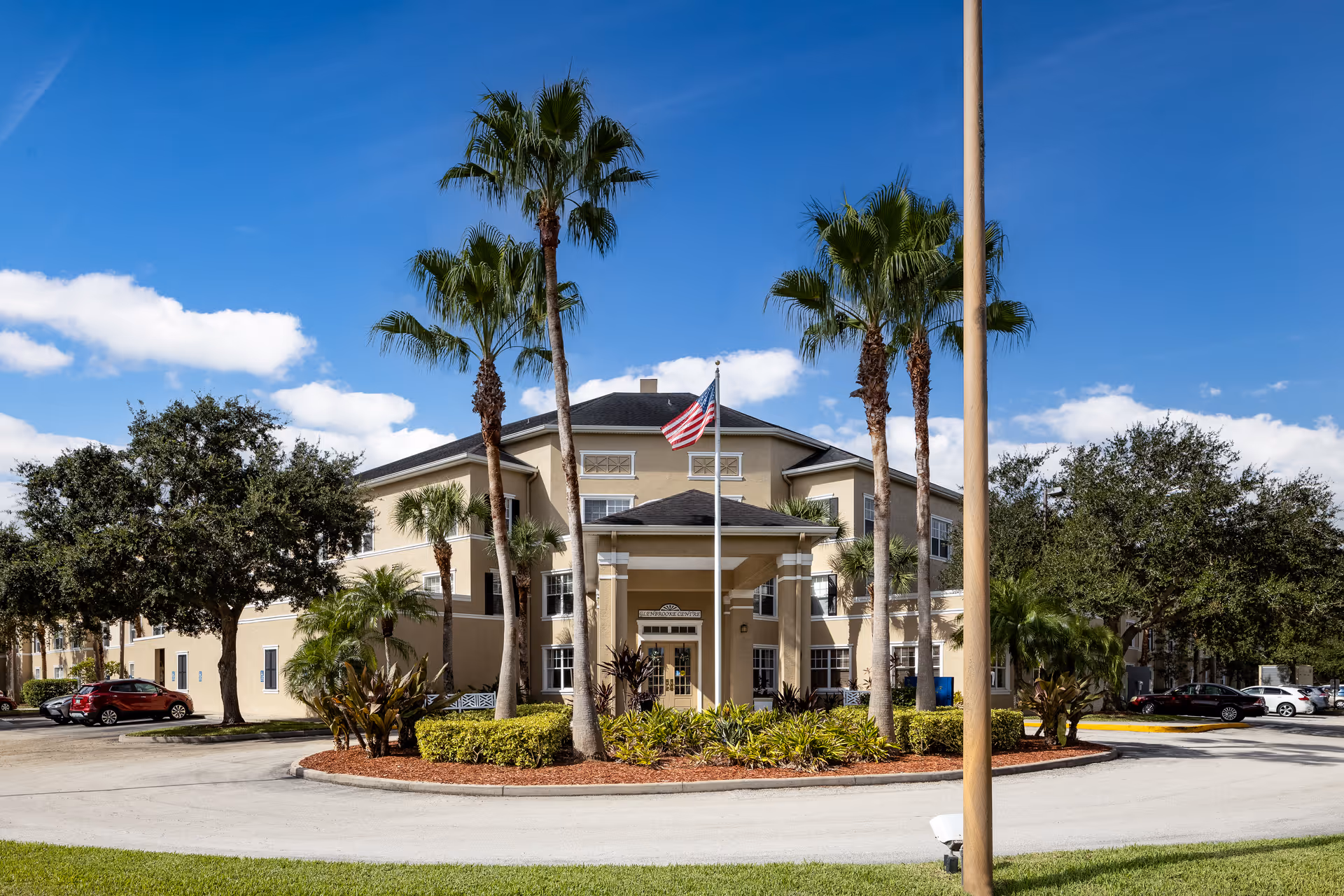Front exterior of a senior living building with palm trees, an American flag, and a circular driveway.