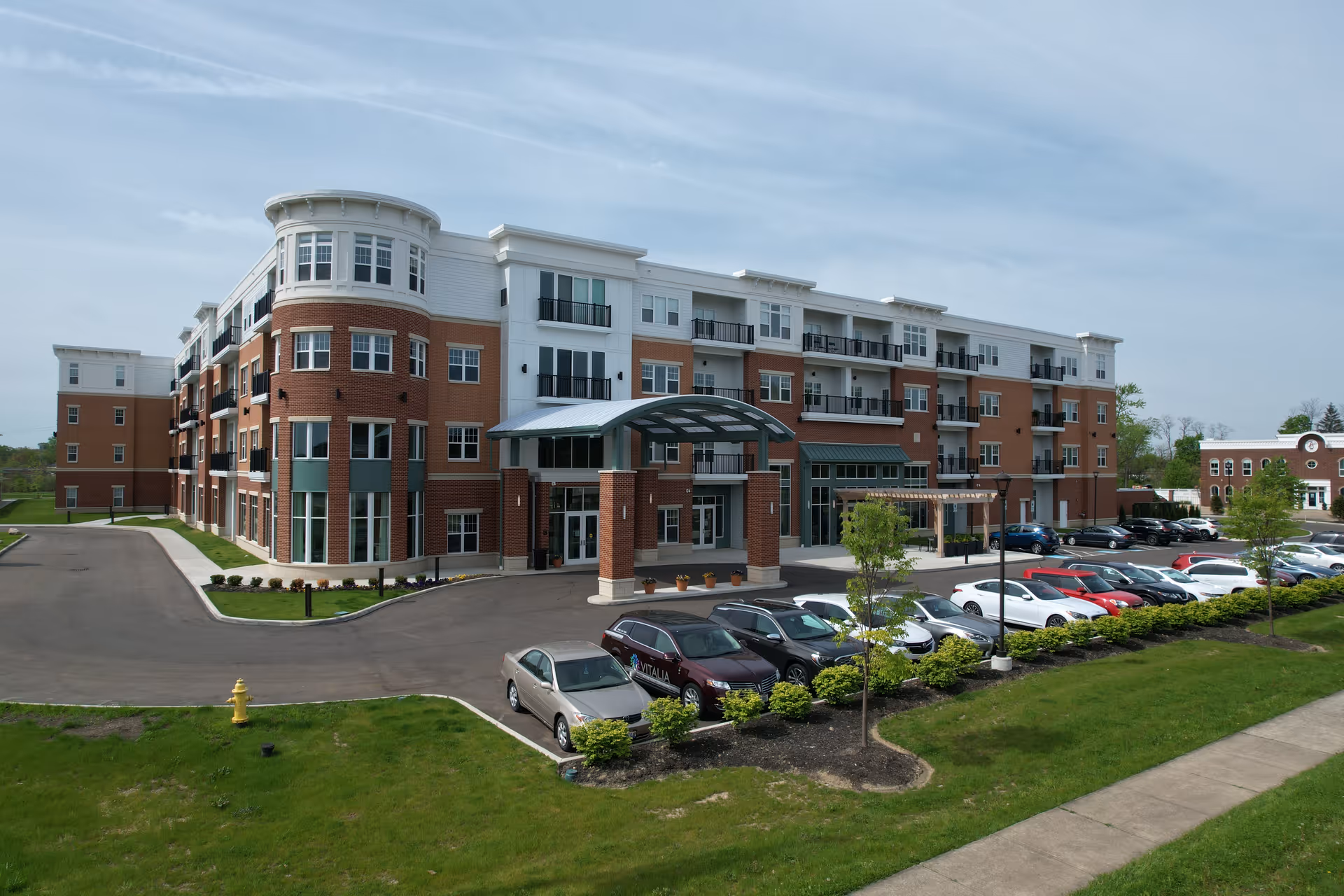 Four-story brick-and-white residential building with a covered main entrance, parked cars, and landscaped grounds.