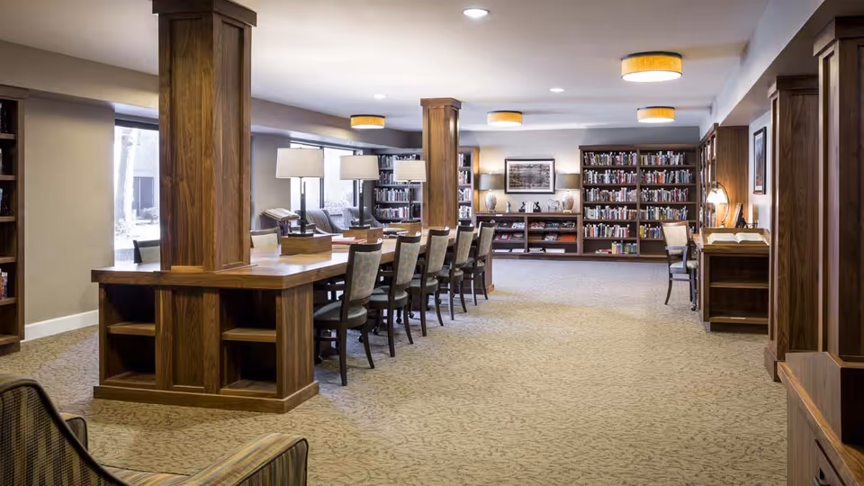 A spacious library or reading room with long wooden tables and chairs arranged in the center. The room features built-in wooden bookshelves filled with books along the walls, several table lamps, and framed artwork. The carpeted floor and warm lighting create a cozy atmosphere.