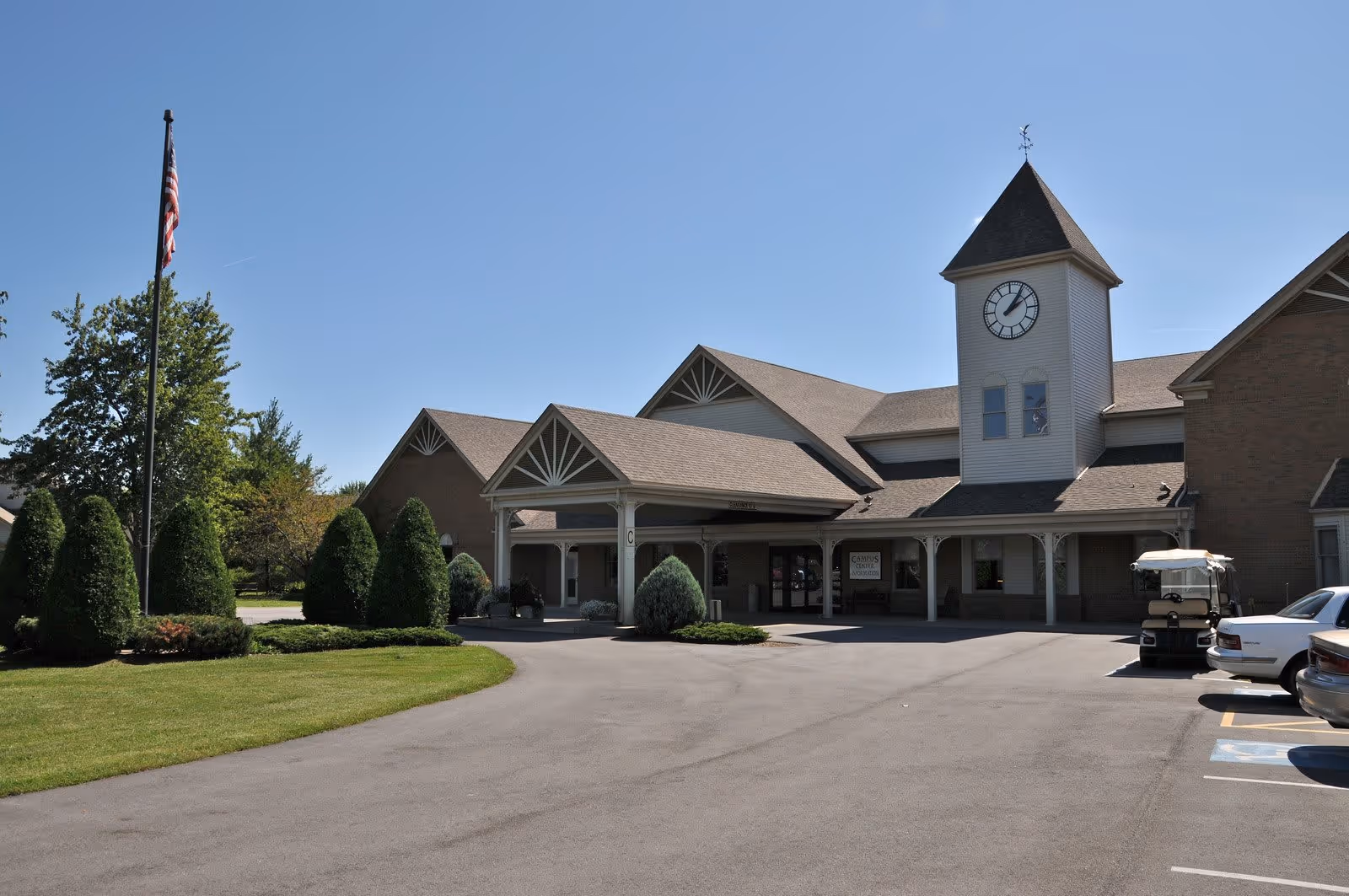 Exterior view of Otterbein St. Marys SeniorLife Community building with a clock tower, covered entrance, neatly trimmed bushes, an American flag on a flagpole, and a parking area with cars and a golf cart.