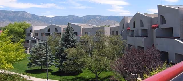 Exterior view of The Montebello on Academy facility showing multiple connected buildings with a modern architectural design, surrounded by green trees and a well-maintained lawn, with mountains visible in the background under a partly cloudy sky.