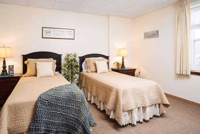 A cozy bedroom with two twin beds, each with beige bedspreads and decorative pillows. There are two wooden nightstands with lamps on either side of the beds. A green plant is placed between the beds, and a window with cream-colored curtains lets in natural light. The walls are light-colored with framed artwork hanging above the beds.