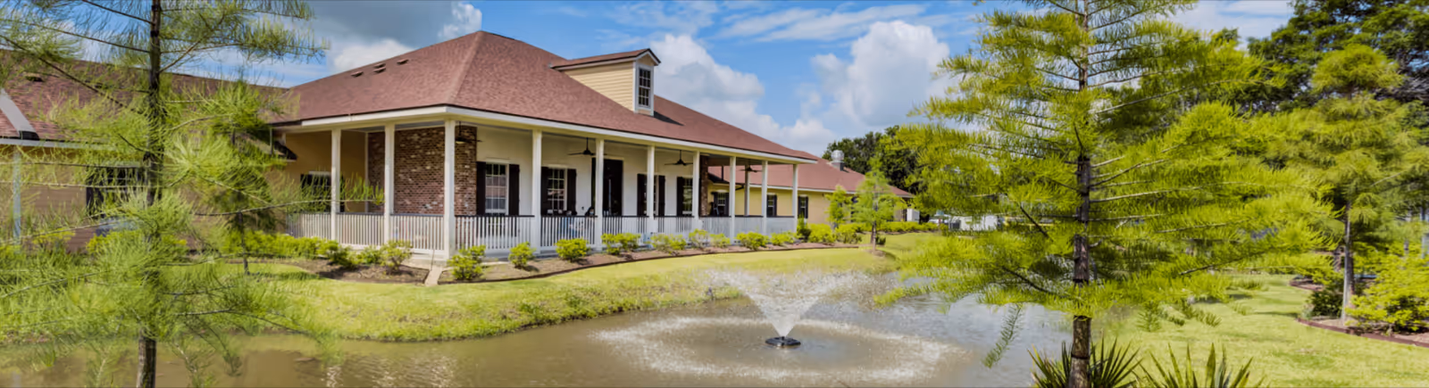 A single-story building with a red roof and a covered porch surrounded by greenery and trees. In front of the building is a pond with a water fountain spraying water upwards under a partly cloudy blue sky.