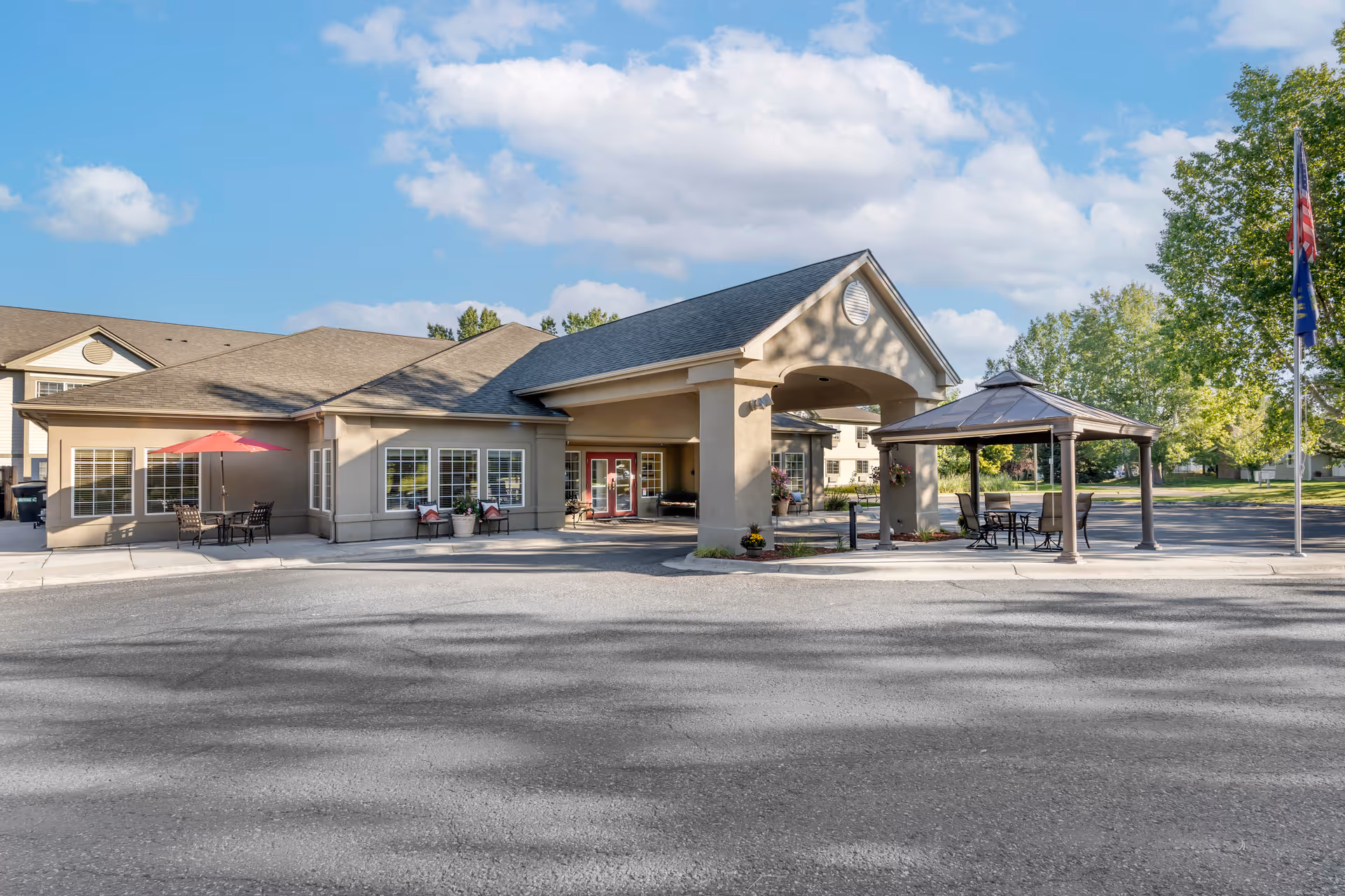 Exterior view of Brookdale Springmeadows facility showing the entrance with a covered driveway, outdoor seating area with tables and chairs under a gazebo, and an umbrella providing shade to another seating area. The building is surrounded by trees and a clear blue sky with some clouds.