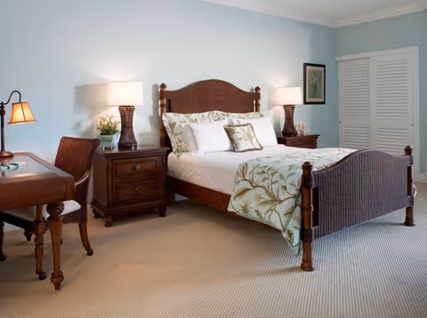 A cozy bedroom with a wooden bed frame featuring a floral bedspread and multiple pillows. There are two matching wooden nightstands on either side of the bed, each with a lamp and decorative items. A wooden desk and chair are positioned to the left, and a closet with white louvered doors is visible on the right. The walls are painted light blue and the floor is carpeted.