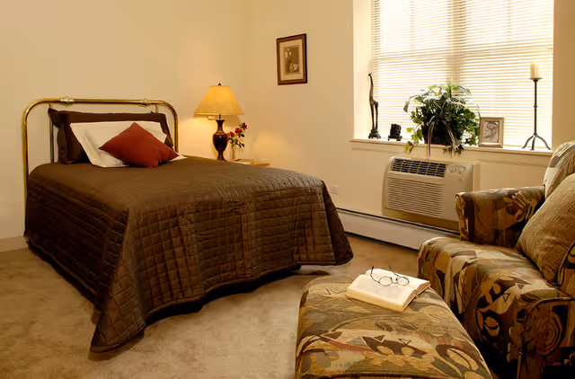 A cozy bedroom with a bed covered in a dark brown quilt and pillows, a bedside table with a lamp and small flower vase, a framed picture on the wall, a window with blinds, an air conditioning unit below the window, and a patterned armchair with a matching ottoman holding an open book and glasses.