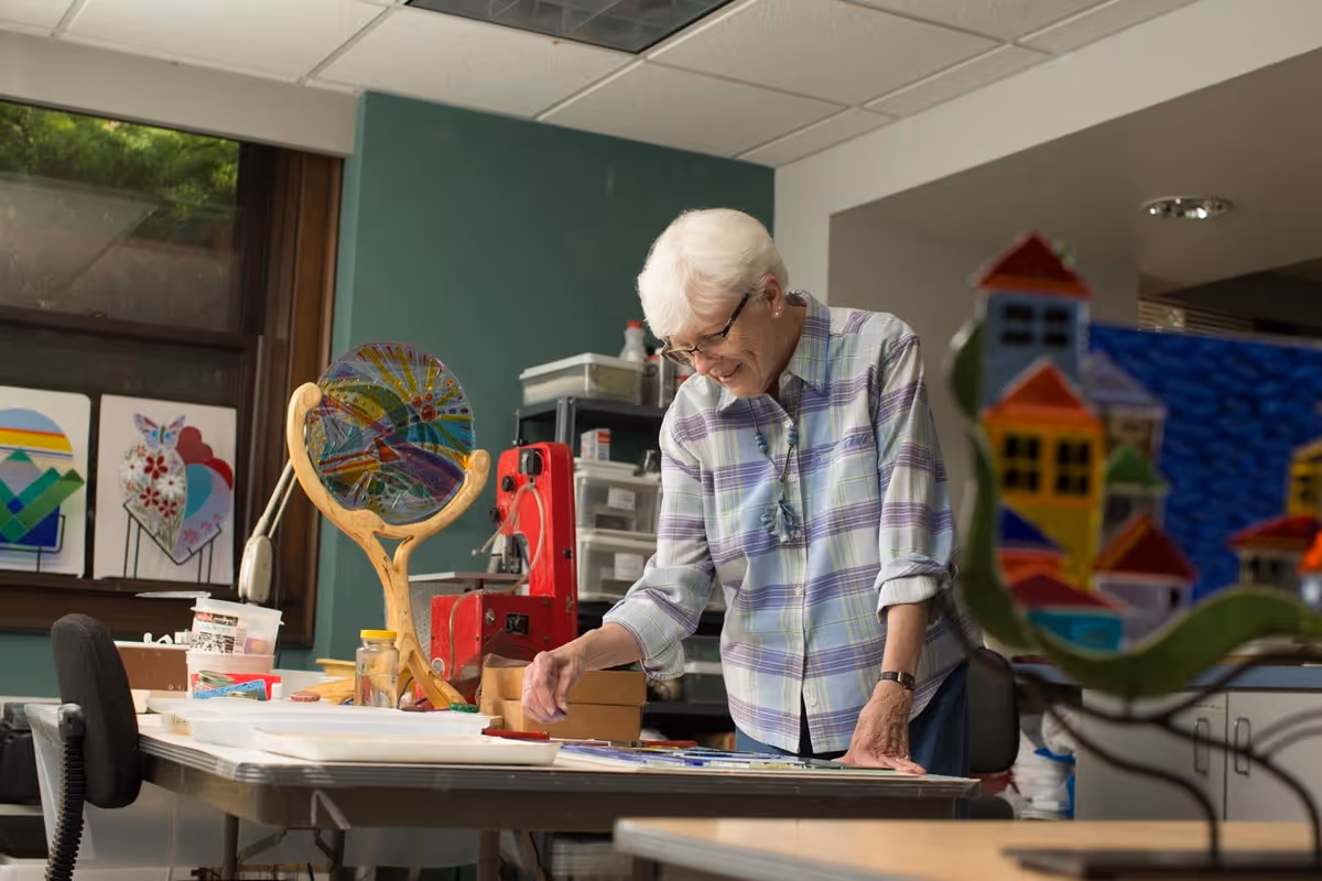 An elderly woman with white hair and glasses is engaged in an art activity at a table in a well-lit room. She is wearing a plaid shirt and smiling while working on a colorful project. The room has art supplies, colorful stained glass pieces, and a window with natural light coming in.