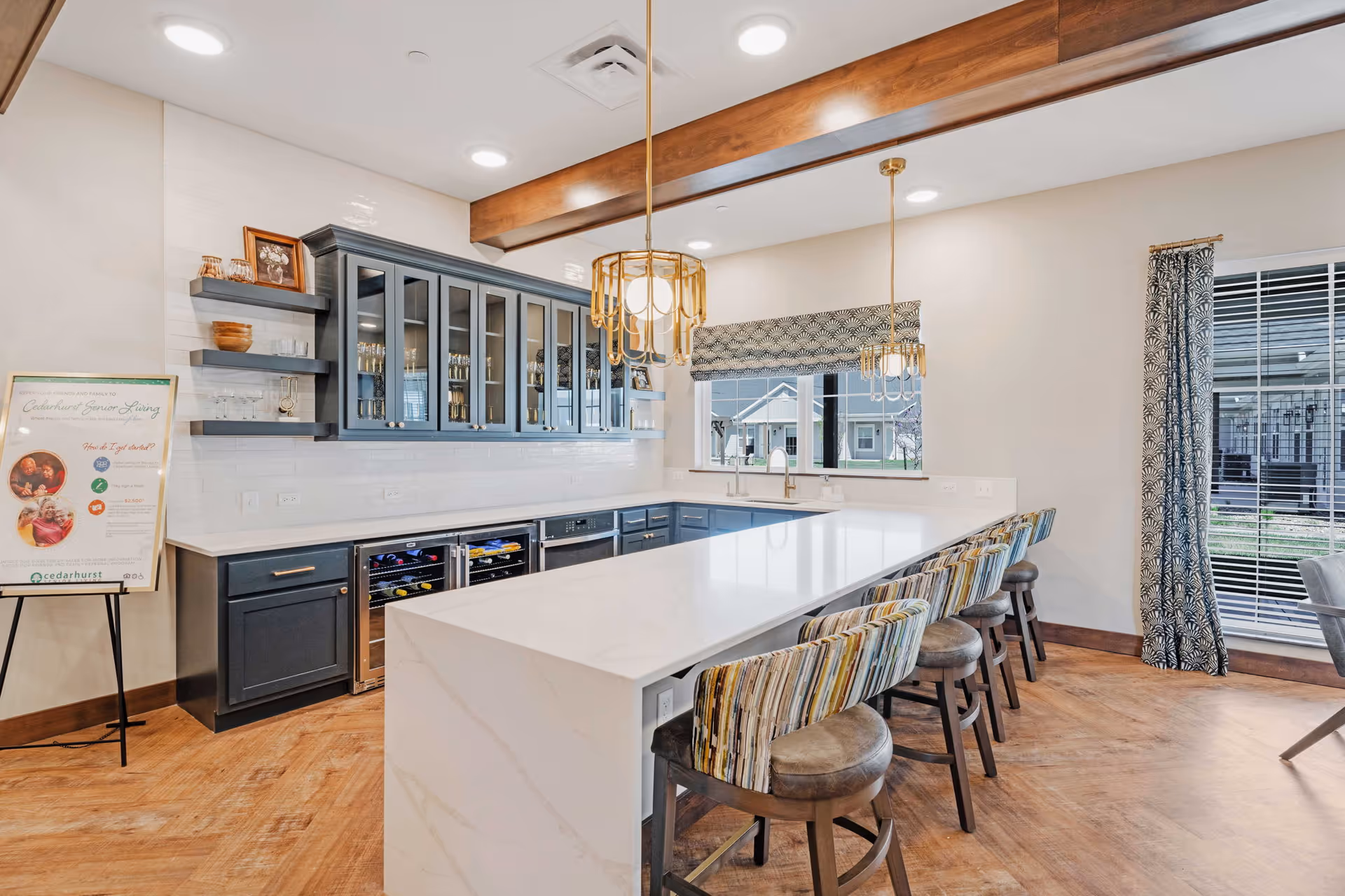 Bright modern kitchen with a long white island, patterned barstools, dark cabinets with glass doors, pendant lights, and large windows.