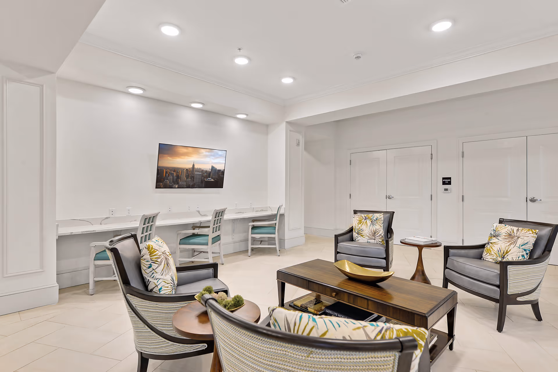 A bright and modern lounge area with four armchairs featuring patterned cushions arranged around a wooden coffee table with a decorative bowl. Behind the seating area, there is a long white desk with three chairs and a wall-mounted TV displaying a cityscape. The room has white walls, recessed lighting, and two closed double doors.