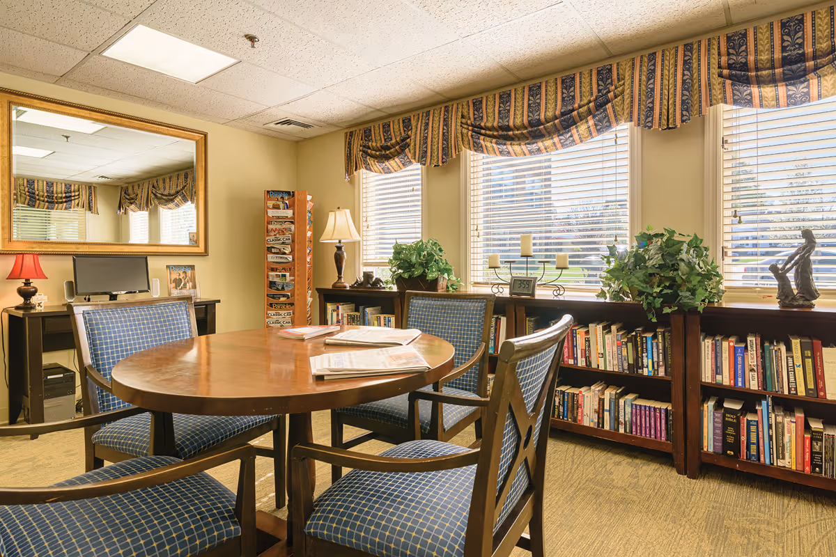 Sunlit common room with a round wooden table and chairs, bookshelves under windows, plants, and lamps.