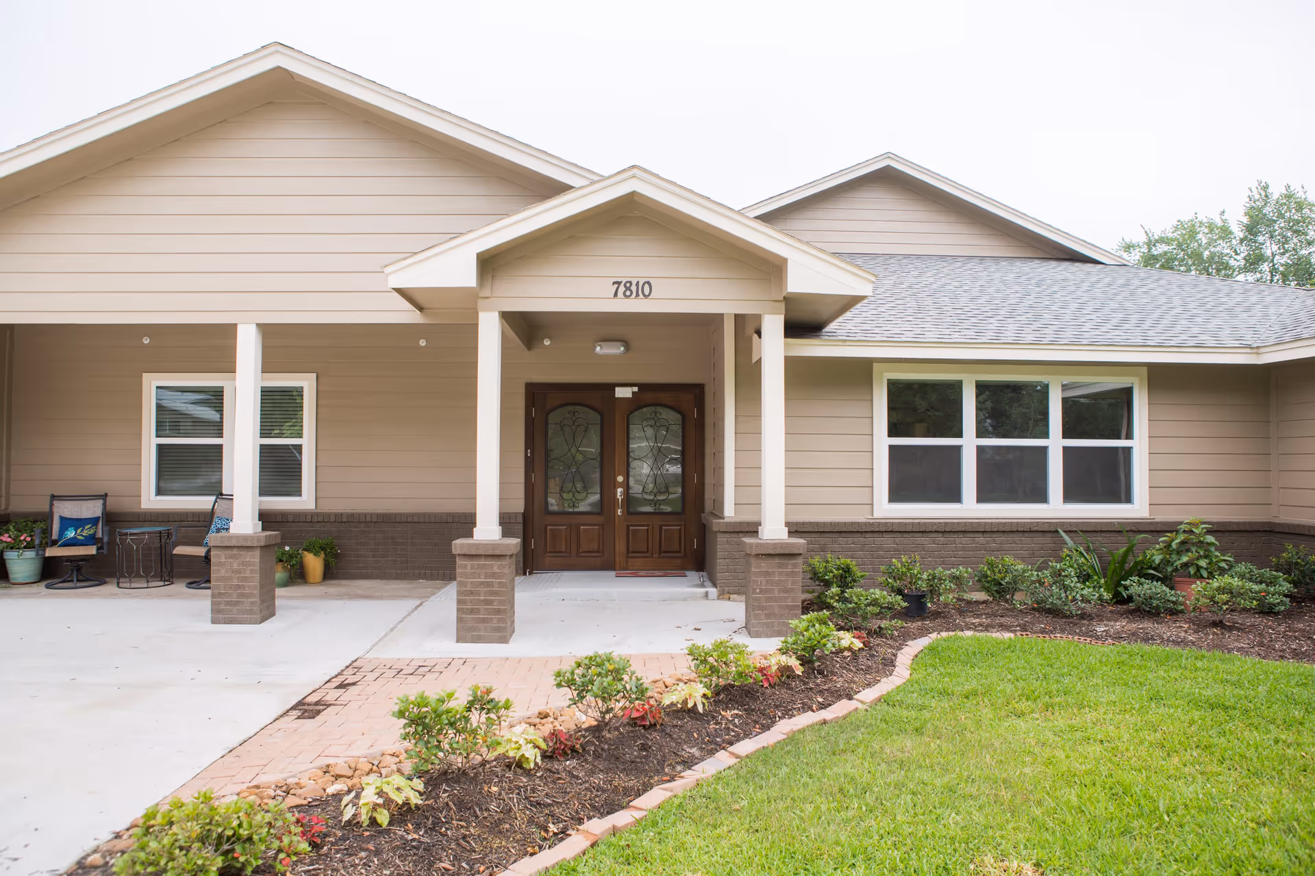 Front exterior view of a single-story building with beige siding and a covered entrance supported by four columns. The entrance has double wooden doors with decorative glass panels. There is a small garden with shrubs and flowers along a brick pathway leading to the entrance. The building number 7810 is displayed above the entrance.