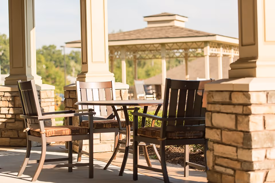 Outdoor seating area with a round table and four wooden chairs with cushions, situated on a covered patio with stone pillars. In the background, there is a gazebo and greenery under a clear sky.