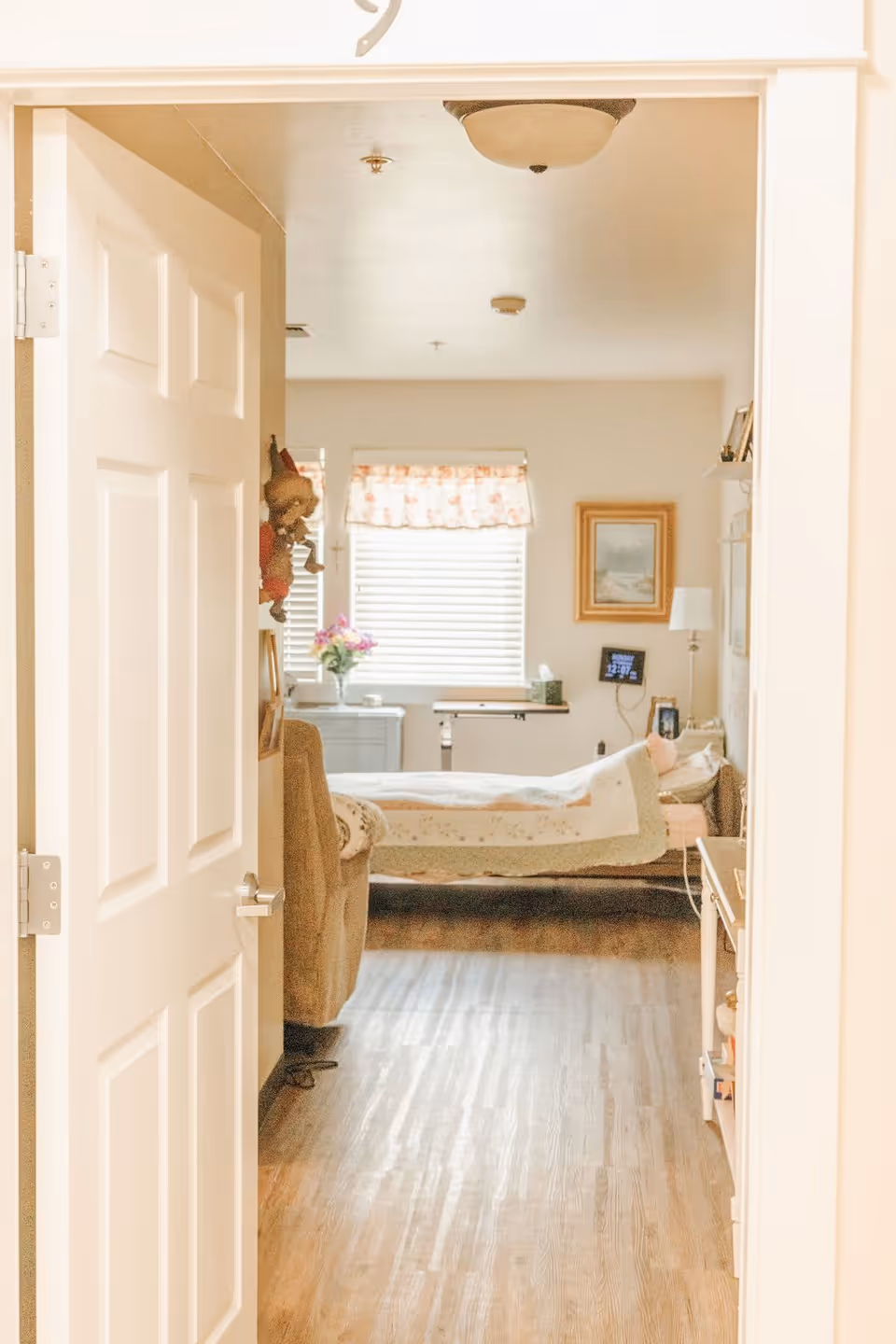 View through an open door into a cozy bedroom with a bed covered in a quilt, a window with floral curtains, a side table with a vase of flowers, a framed picture on the wall, and a floor lamp. The room has wooden flooring and soft lighting.