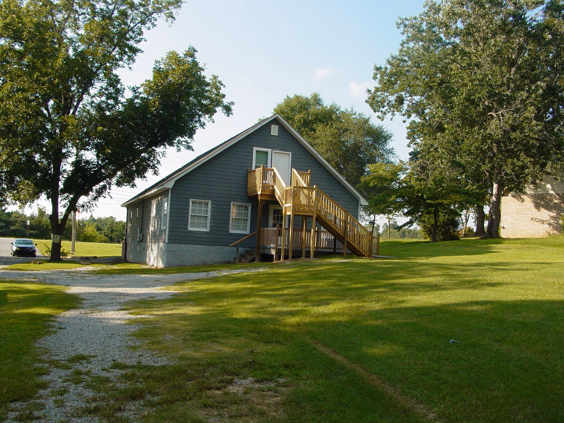 A small gray building with white trim and a wooden staircase leading to a second-floor door. The building is surrounded by green grass and large trees under a clear blue sky. A gravel driveway leads up to the building, and a parked car is visible in the background.