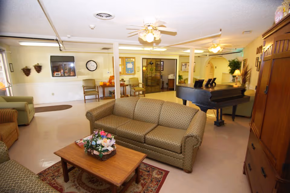 A spacious living room area in a nursing facility featuring a patterned sofa, armchairs, a wooden coffee table with a floral arrangement, a grand piano, ceiling fans with lights, and various decorative elements including wall clocks and plants.