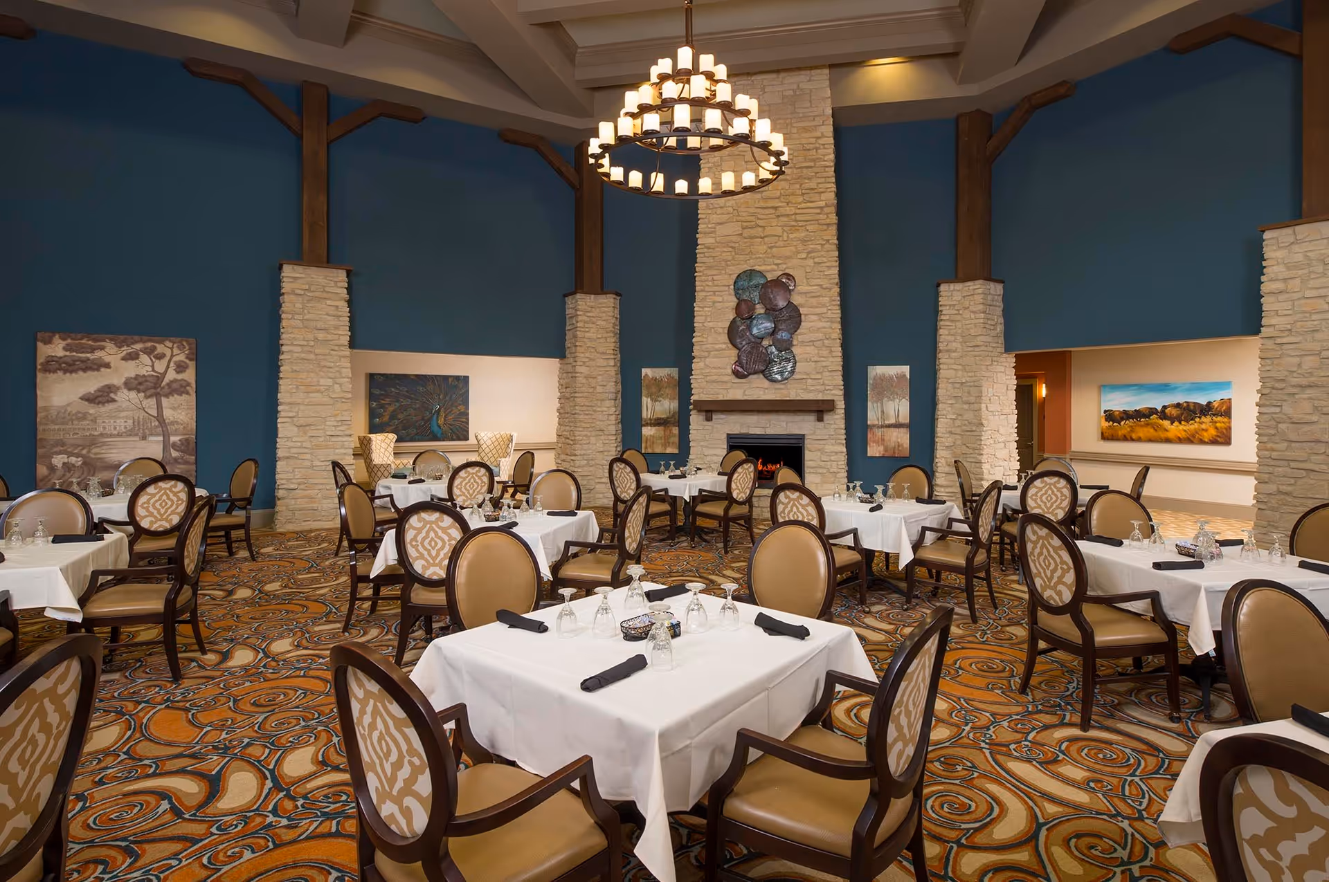 A spacious dining room with multiple tables covered in white tablecloths, each set with glasses and black napkins. The room features a patterned carpet, stone pillars, blue walls, and a large chandelier hanging from a beamed ceiling. There is a stone fireplace with decorative art above it and framed paintings on the walls.