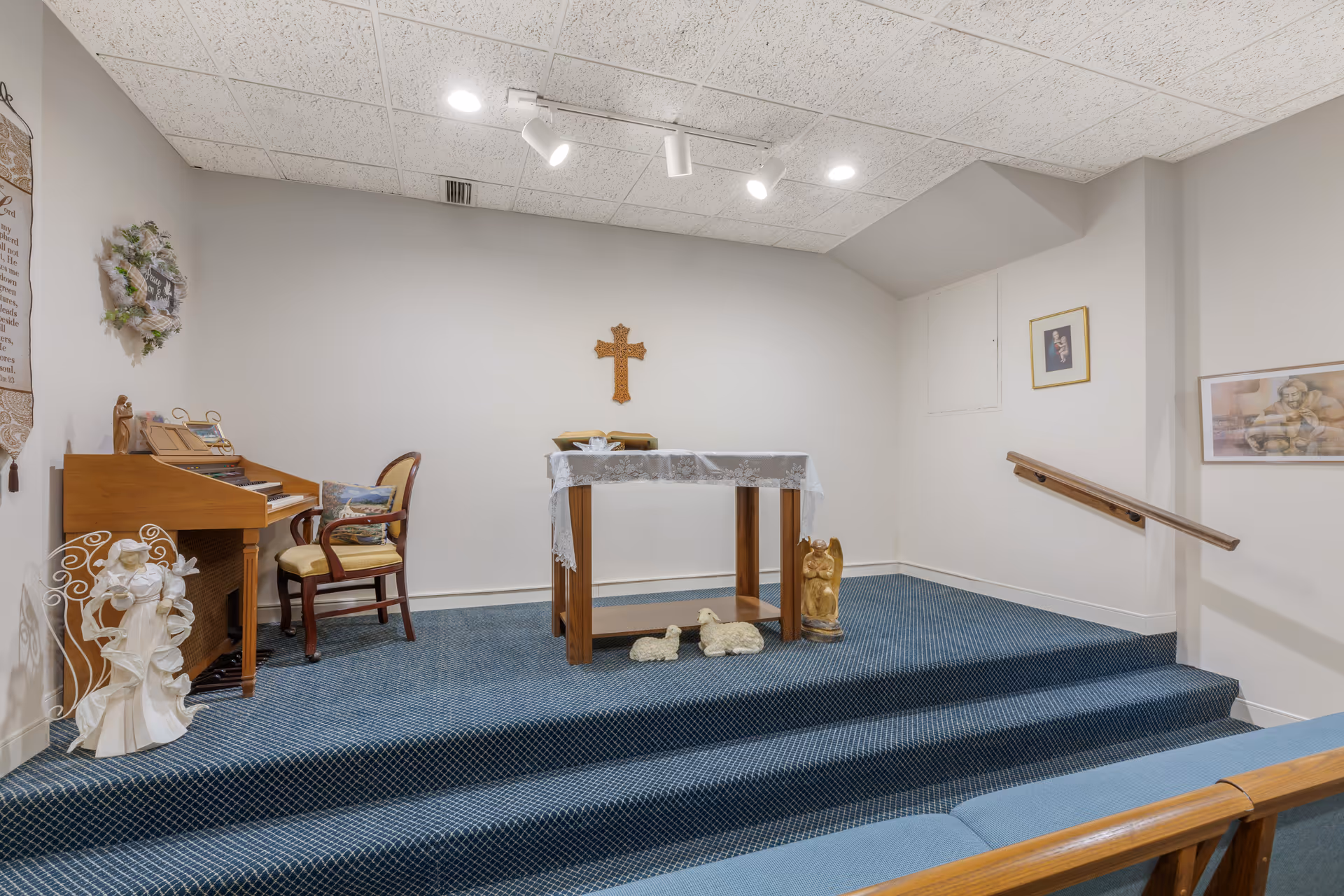 Small interior chapel with an altar draped in a cloth, a wooden organ and chairs, religious statues, and a cross on the wall.