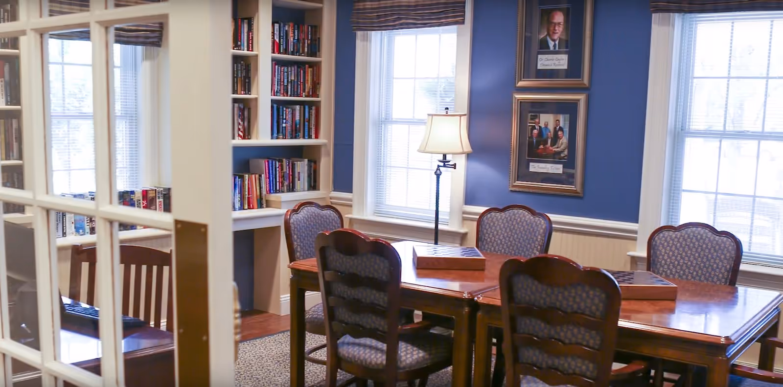 A cozy room with a wooden table surrounded by six upholstered chairs, two large windows with blinds, a floor lamp, built-in bookshelves filled with books, and framed portraits on a blue wall.