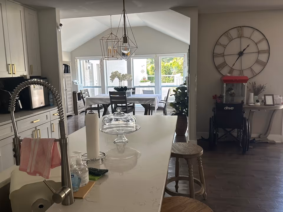 Interior view of a senior living facility showing a modern kitchen island with a sink, soap dispensers, paper towel holder, and a glass cake stand. In the background, there is a dining area with a table covered by a white tablecloth and chairs, large windows letting in natural light, a large decorative wall clock, a wheelchair, a popcorn machine, and a small table with decorative items.