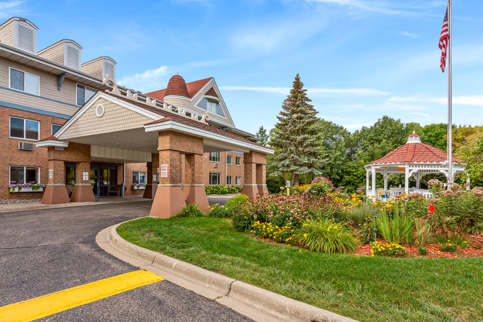 Entrance of a senior living facility with a covered drop-off area, a well-maintained garden with colorful flowers, a white gazebo with a red roof, and an American flag on a flagpole under a blue sky.