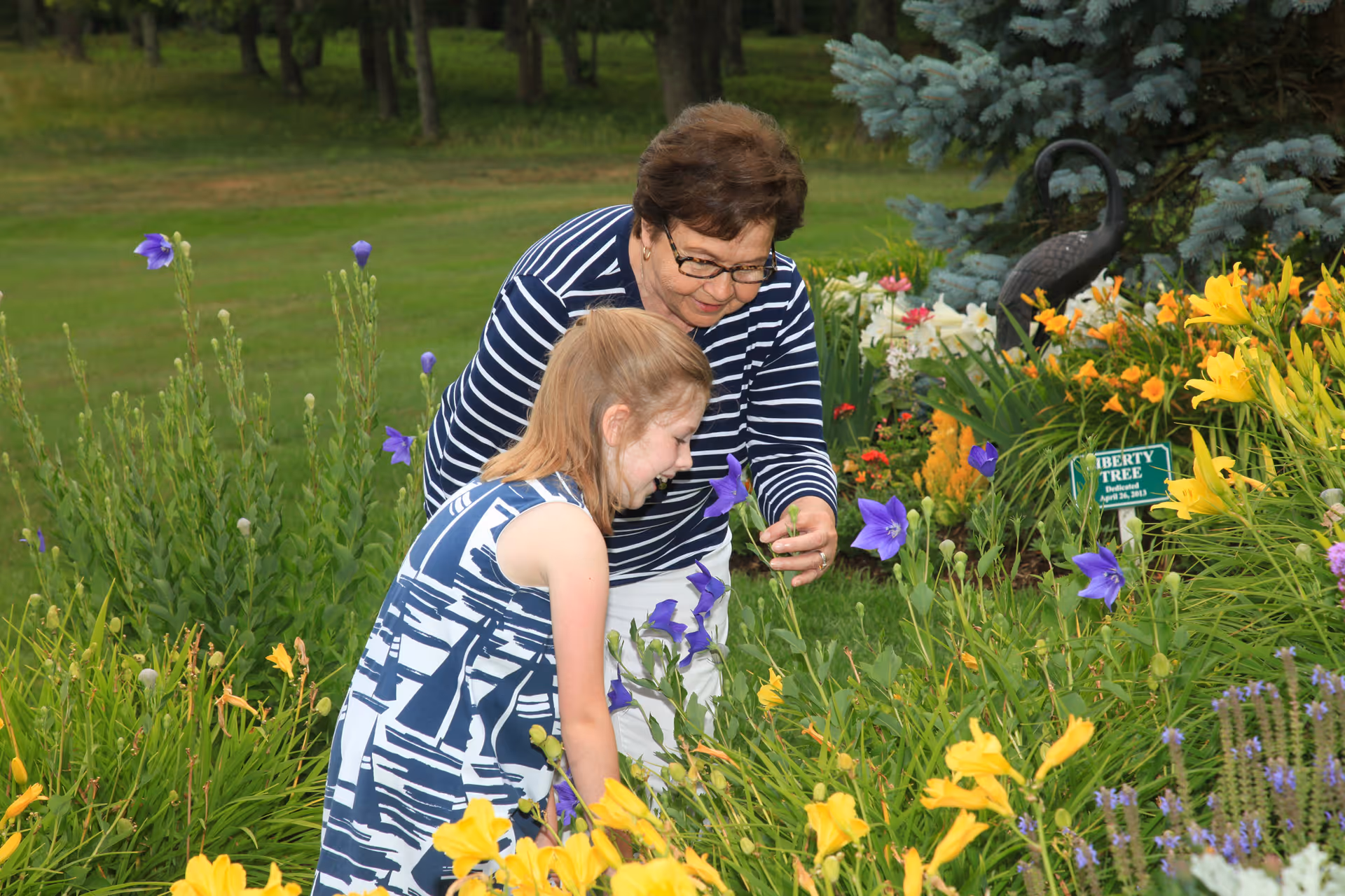 An older woman and a young girl are in a garden surrounded by colorful flowers including yellow lilies and purple blooms. The woman is wearing glasses and a striped shirt, and the girl is wearing a blue and white patterned dress. They are both looking closely at the flowers, with the woman gently holding one of the purple flowers.
