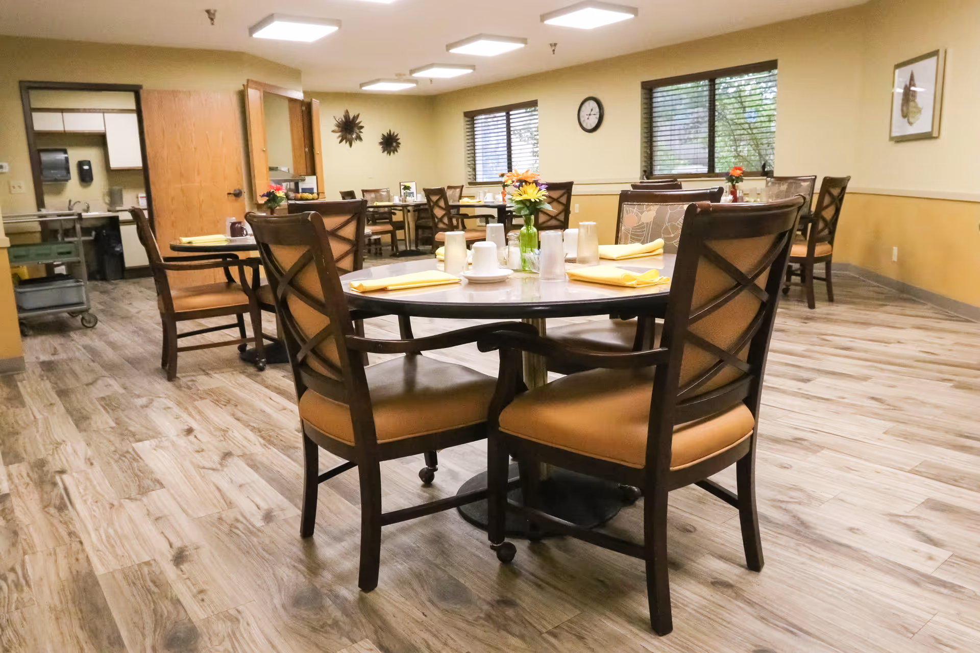 Dining room with round tables set with yellow napkins and small flower arrangements, surrounded by wooden chairs on wood-look flooring.