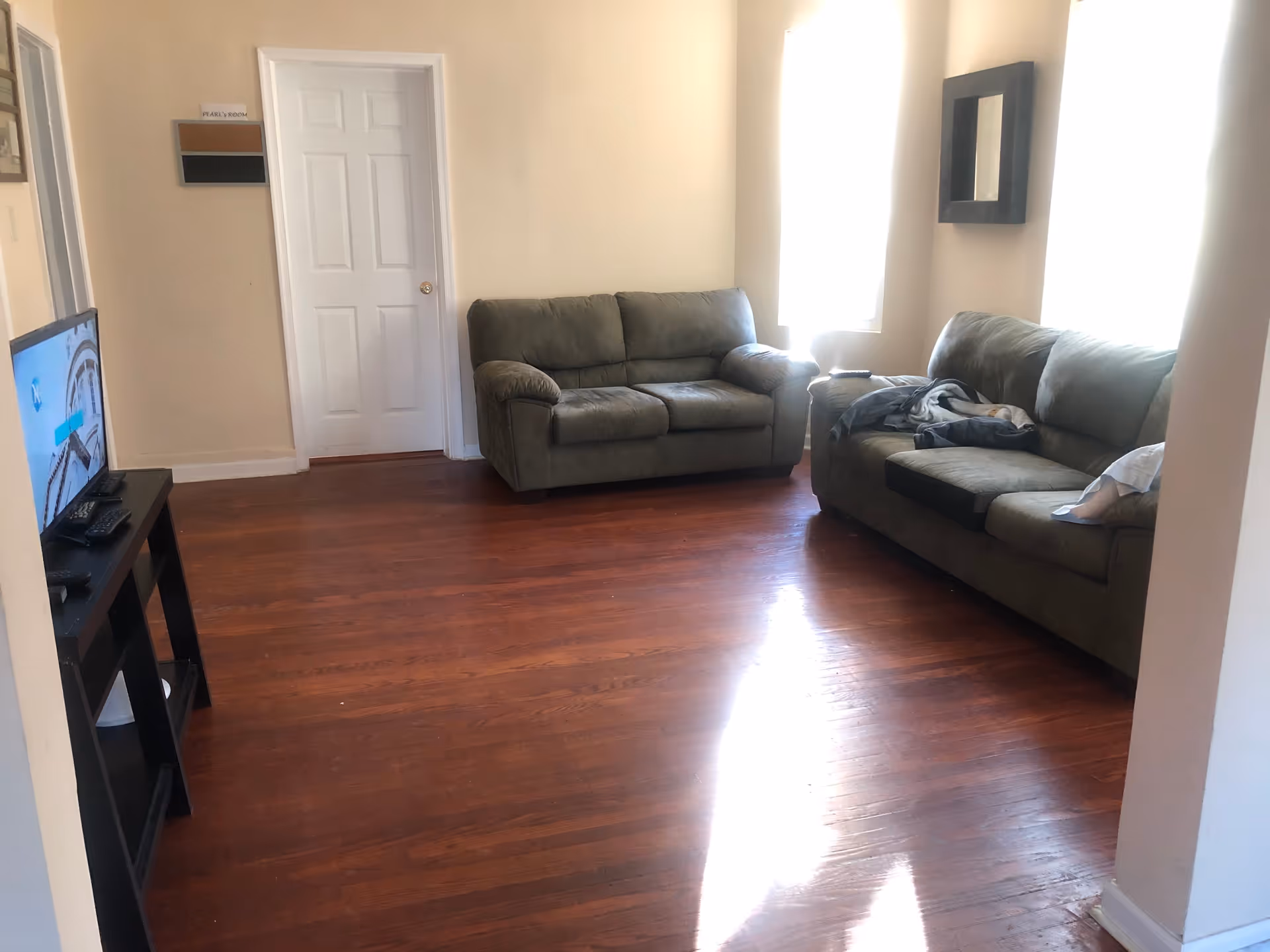 A living room with two green upholstered sofas, a flat-screen TV on a black stand, hardwood floors, and two windows with bright sunlight streaming in. There is a door in the background and a small wall-mounted mirror between the windows.