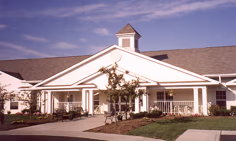 Front exterior view of a single-story senior living community building with a white facade, a peaked roof with a small cupola, a covered porch with columns, benches, and landscaped greenery including small trees and shrubs.