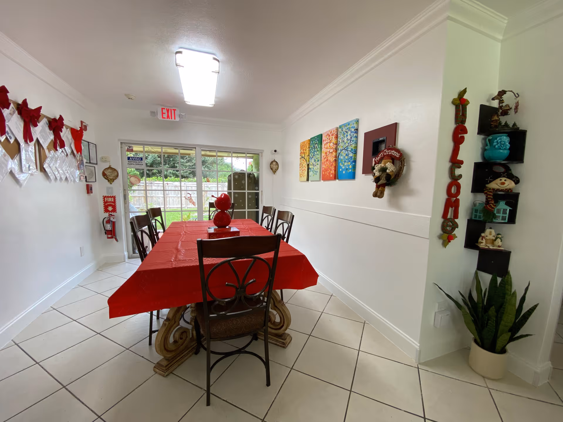 A dining room with a long table covered with a red tablecloth and surrounded by chairs. The room has white tiled floors and white walls decorated with colorful paintings and holiday decorations. A glass sliding door at the end of the room opens to a green outdoor area. There is a fire extinguisher mounted on the left wall and a 'Welcome' sign with decorative shelves on the right wall.