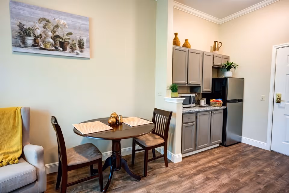 A small dining area with a round wooden table set with two placemats and two chairs. To the right is a compact kitchen with gray cabinets, a microwave, a small refrigerator, and decorative plants and vases on top of the cabinets. A beige armchair with a yellow throw blanket is partially visible on the left side. The floor is wood, and the walls are light-colored with a painting of potted plants hanging above the dining table.