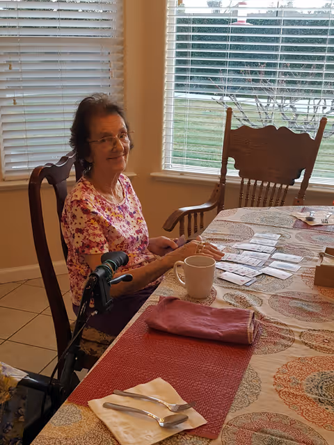 An elderly woman with glasses sitting at a dining table with a walker beside her. She is smiling and wearing a floral shirt. The table is covered with a patterned tablecloth and has a white mug, a folded cloth napkin, and several cards spread out on it. There are large windows with blinds in the background showing a green outdoor area.
